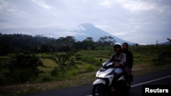 Orang mengendarai sepeda motor melewati dengan latar belakang Gunung Agung, gunung berapi berstatus "awas", di Kabupaten Karangasem, Bali, Indonesia, 24 September 2017.