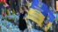 Una mujer lleva una bandera con los colores nacionales de Ucrania y el retrato de un militar ucraniano caído en un monumento improvisado en la Plaza de la Independencia en Kiev, el 2 de noviembre de 2024, en medio de la invasión rusa en Ucrania. (Foto de Sergei SUPINSKY / AFP)