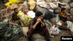 FILE - Afghan refugees sit on sacks filled with used plastic items to sell at their makeshift shelter in a slum on the outskirt of Lahore, Jan. 12, 2015.