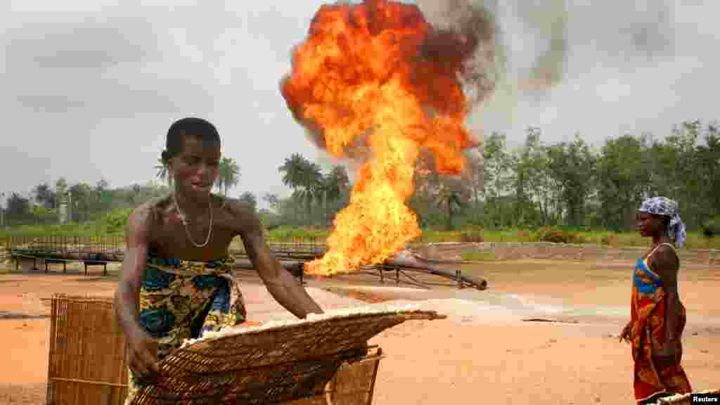 A woman dries a basket of cassava beside flames from an oil flow station.