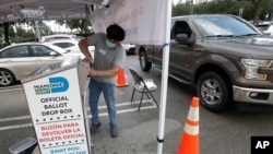 A poll worker wearing personal protective equipment deposits a Vote-by-Mail ballot for a voter, Aug. 3, 2020, at a poling station at the Miami-Dade County Elections Department in Miami. 