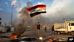 Un manifestante ondea una bandera iraquí mientras otros bloquean las calles con fuego cerca de la plaza Tahrir en Bagdad, Irak, el miércoles, 8 de enero de 2020.