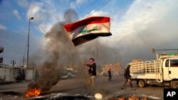 Seorang pengunjuk rasa mengibarkan bendera Irak sementara demonstran membakar jalan-jalan dekat Tahrir Square selama demonstrasi untuk memprotes serangan rudal Iran, di Baghdad, Irak, Rabu, 8 Januari 2020. (Foto: AP)