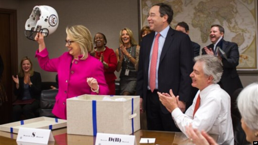 Secretary of State Hillary Rodham Clinton holding up a football helmet presented to her as she returned to work after a month-long absence caused first by a stomach virus, then a fall and a concussion, and finally a brief hospitalization for a blot clot n