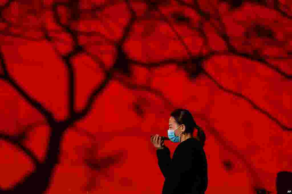 A woman wearing a face mask to help curb the spread of the coronavirus talks on her smartphone as she walks by tree shadow cast on a truck parked on a street in Beijing.