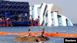 Bañistas se divierten frente a los restos del Costa Concordia, al que se le sumergido rodeado por grúas.