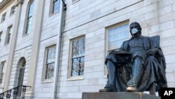 FILE - The John Harvard statue at Harvard University, a popular tourist attraction at the campus in Cambridge, Masssachusetts, sits adorned with a mask, March 14, 2020. The university is restricting spending in the wake of the coronavirus outbreak.