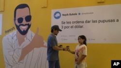 Una pareja se encuentra frente a un mural político del presidente Nayib Bukele, el mensaje: "Ordeno que vendan 3 pupusas por un dólar", parte de una ofensiva del gobierno para bajar los precios de los alimentos, en San Salvador, El Salvador, el viernes 12 de julio de 2024.