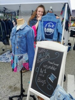 Ellie Heath stands next to some of her designs - repurposed jeans jackets adorned with her distinctive artistic touch (Photo: F. Elmasry / VOA)