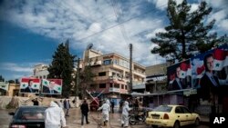 FILE - In this March 24, 2020, file photo, workers spray disinfectant to prevent the spread of the coronavirus on a street lined with billboards showing Syrian President Bashar Assad, in Qamishli, Syria.
