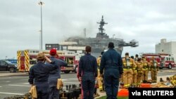 Marineros de la Armada de EE.UU. saludan a la bandera la mañana del 14 de julio de 2020, detrás del buque de asalto anfibio USS Bonhomme Richard, que se incendió dos días antes en la Base Naval de San Diego, California.