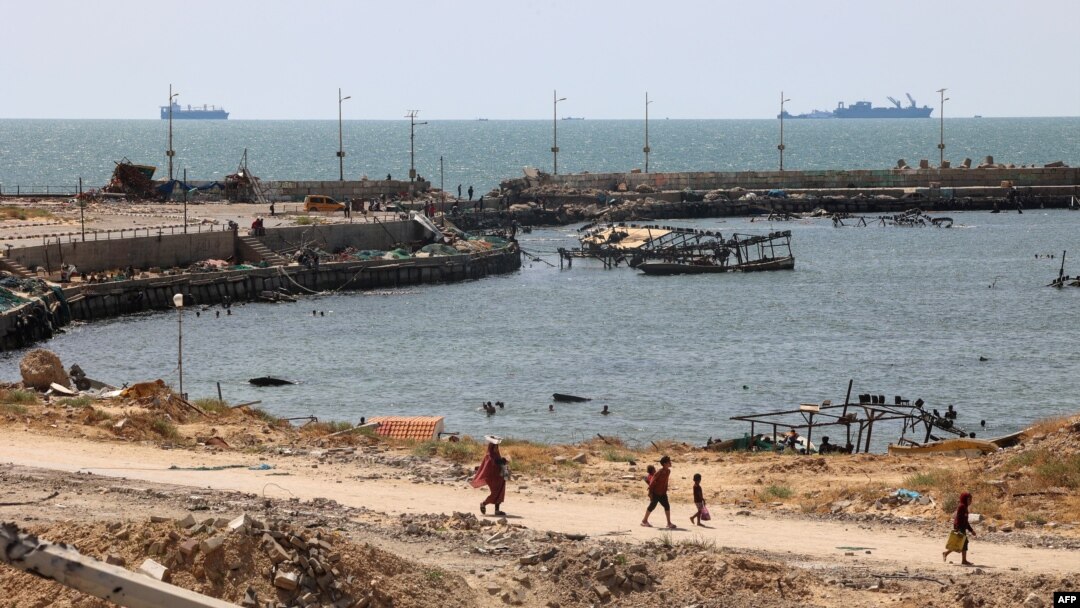 Palestinians walk past a jetty in Gaza City with a view of navy vessels off the coast as part of a humanitarian "maritime corridor" announced by U.S. Central Command on May 17, 2024.