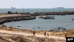 Palestinians walk past a jetty in Gaza City with a view of navy vessels off the coast as part of a humanitarian "maritime corridor" announced by U.S. Central Command on May 17, 2024.