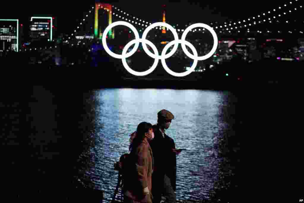 A man and a woman walk past the Olympic rings hanging above the water in the Odaiba section in Tokyo, Japan.