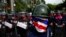 FILE - An anti-government protester wearing a mask painted in the colors of the Thai national flag looks on as riot police officers stand guard outside the parliament in Bangkok, Aug. 7, 2013. 