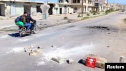 FILE - Men ride a motorbike past a hazard sign at a site hit by an airstrike on Tuesday in the town of Khan Sheikhoun in rebel-held Idlib, Syria, April 5, 2017. The hazard sign reads, "Danger, unexploded ammunition." 