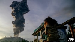 A woman and child look on at volcanic ash rising into the air during the eruption of Mount Ibu, as seen from Duono village, North Maluku province, Jan. 15, 2025. 