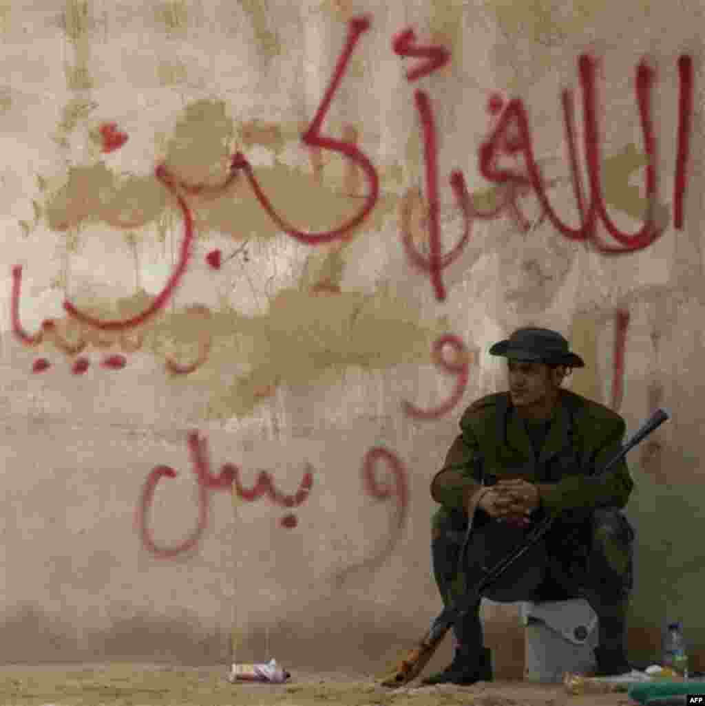 A Libyan rebel sits under a Arabic slogan reading " god is great, only Libya' on the frontline of the outskirts of the city of Ajdabiya, south of Benghazi, eastern Libya, Monday, March 21, 2011. The international military intervention in Libya is likely t