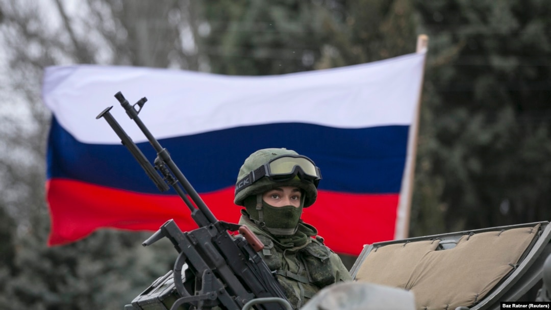 Seorang pria pro-Rusia (tidak terlihat) memegang bendera Rusia di belakang seorang prajurit bersenjata di atas kendaraan tentara Rusia di luar pos penjaga perbatasan Ukraina di kota Balaclava, Krimea, 1 Maret 2014. (Foto: REUTERS/Baz Ratner)