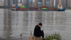 In this Monday, April 13, 2020, photo, a man wearing a mask against the new coronavirus checks his phone as a container ship cruises along the Yangtze River in Wuhan in Central China's Hubei province. China's exports fell further in March compared…