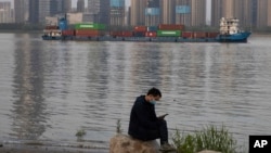 In this Monday, April 13, 2020, photo, a man wearing a mask against the new coronavirus checks his phone as a container ship cruises along the Yangtze River in Wuhan in Central China's Hubei province. China's exports fell further in March compared…