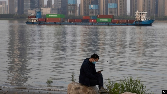 In this Monday, April 13, 2020, photo, a man wearing a mask against the new coronavirus checks his phone as a container ship cruises along the Yangtze River in Wuhan in Central China's Hubei province. China's exports fell further in March compared…