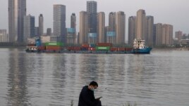 In this Monday, April 13, 2020, photo, a man wearing a mask against the new coronavirus checks his phone as a container ship cruises along the Yangtze River in Wuhan in Central China's Hubei province. China's exports fell further in March compared…