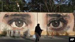 FILE - An Afghan woman waits for transportation in front street art on a barrier wall of the NDS (National Directorate of Security) in Kabul, Afghanistan. 