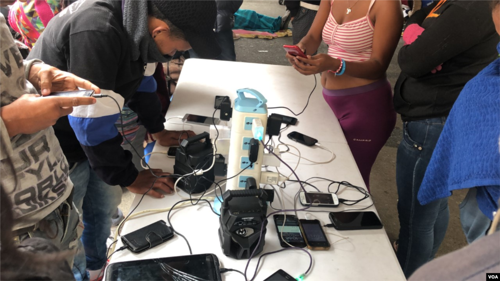 Los inmigrantes venezolanos se reúnen en la mañana alrededor de una mesa donde la Cruz Roja de Colombia proporciona enchufes eléctricos donde pueden cargar sus teléfonos celulares. Ipiales, Colombia, 11 de septiembre de 2019. Foto: Celia Mendoza - VOA. 