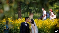 FILE - People wearing face masks pass by newlyweds kissing as they pose for wedding photos at the Olympic Forest Park in Beijing on July 2, 2020. 