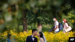 FILE - People wearing face masks pass by newlyweds kissing as they pose for wedding photos at the Olympic Forest Park in Beijing on July 2, 2020. 