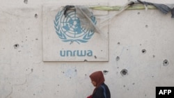 FILE - Seorang perempuan Palestina berjalan melewati tembok yang rusak dengan logo UNRWA di kamp pengungsi di Rafah, Jalur Gaza selatan, 28 Mei 2024, di tengah konflik Israel-Hamas. (Eyad BABA / AFP)