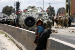 A woman carries a child as members of the security forces stand guard during a protest in Senkata, El Alto, Bolivia, Nov. 19, 2019.