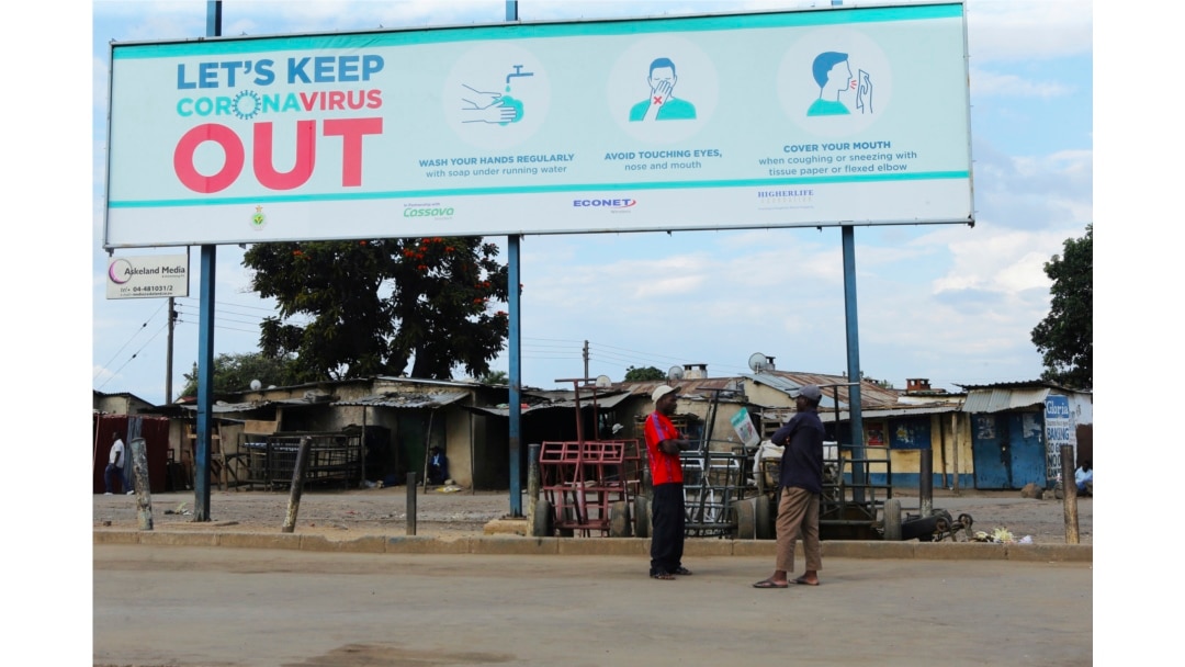FILE - Two men stand underneath a giant poster showing advice on how to prevent the spread of the coronavirus, during a lockdown to control the virus, in Harare, Zimbabwe, April, 5, 2020.