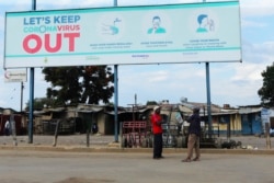 FILE - Two men stand underneath a giant poster showing advice on how to prevent the spread of the coronavirus, during a lockdown to control the virus, in Harare, Zimbabwe, April, 5, 2020.