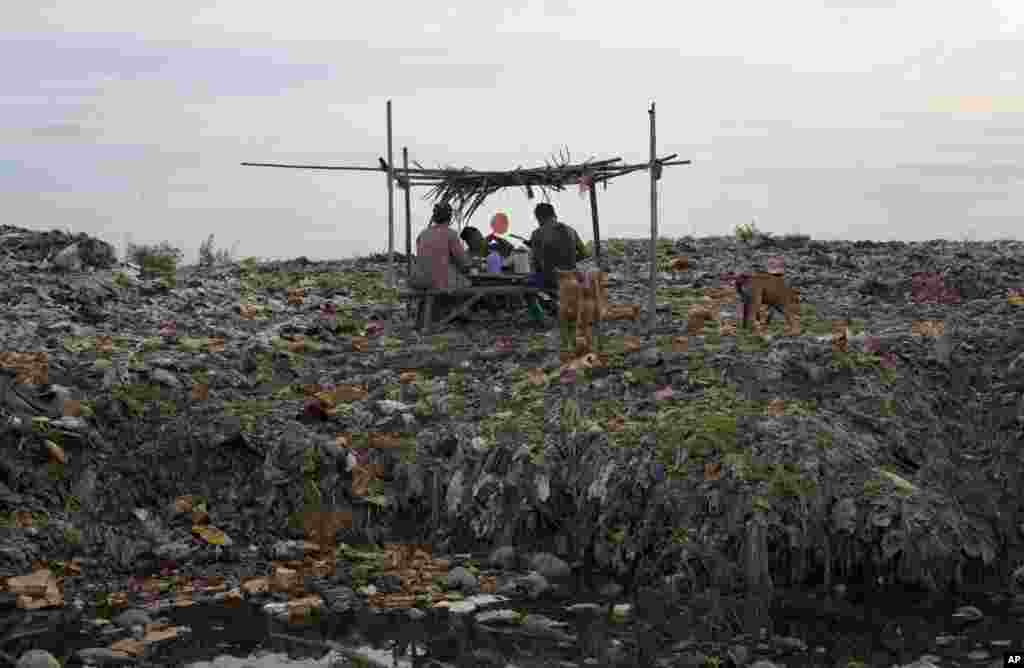 A makeshift tea shop owner sits with garbage sorters on a pile of trash while using a hand held fan to keep bugs away at a suburban garbage dump north of Rangoon, Burma.