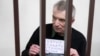 Imprisoned Kremlin critic Alexei Gorinov stands in a cage at the court holding a banner saying "Stop killing, stop the war" in Russian, at his second trial for criticizing Russia's actions in Ukraine, in Vladimir, Russia, Nov. 29, 2024. 