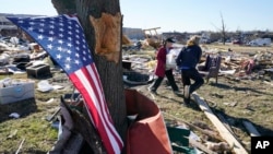 A U.S. flag hangs from a tree as two women carry items recovered from tornado wreckage in Mayfield, Kentucky, Dec. 12, 2021. 