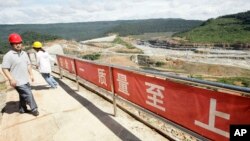 In this Oct. 6, 2012 photo, a Chinese engineer, left, walks by a fence with Chinese slogans reading: Safety first, highest quality and quantity near an entrance of a dam construction site by China National Heavy Machinery Corporation on the Tatay River in Koh Kong province, some 210 kilometers (130 miles) west of Phnom Penh.