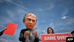 Seorang pria mengenakan topeng Presiden FIFA Sepp Blatter memegang uang Francs Swiss berdiri di sebelah perempuan memegang poster bertuliskan "Game over for Blatter" dalam sebuah demo di depan Hallenstadium tempat Kongres FIFA keCongress takes place in Zurich,