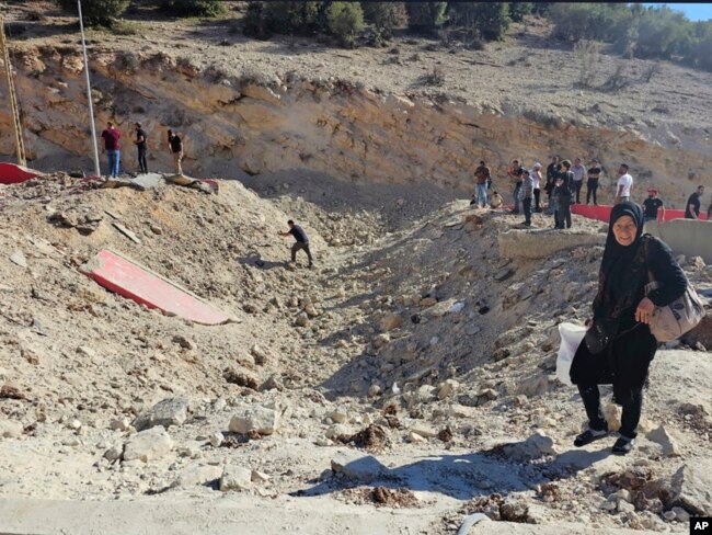 Seorang perempuan membawa barang bawaan menyeberang ke Suriah dengan berjalan kaki melewati kawah akibat serangan Israel yang memotong jalan antara Lebanon dan Suriah di penyeberangan Masnaa, Lebanon, Jumat, 4 Oktober 2024. (Foto: Samer Husseini/AP Photo)