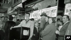 FILE - Phil Bustos of Trinidad, Colorado, displays a television set he bought for $29 at a Washington's Birthday sale in Washington, D.C., Feb. 22, 1954. Other bargain hunters waited to get into the packed store during the annual holiday sale.