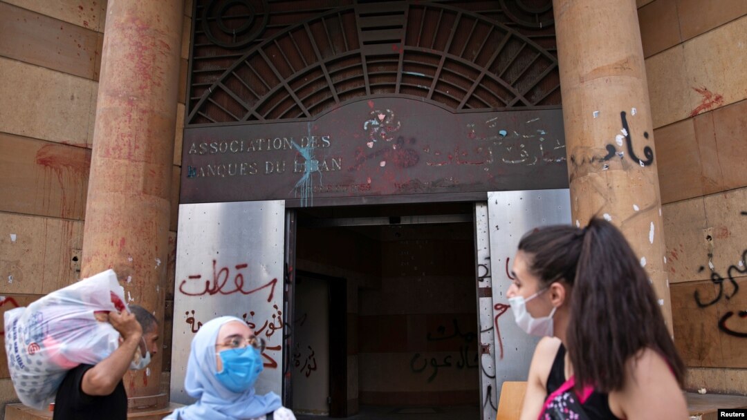 FILE - People wearing face masks stand outside the damaged entrance of the Association of Banks, in Beirut, Lebanon, Aug. 21, 2020.