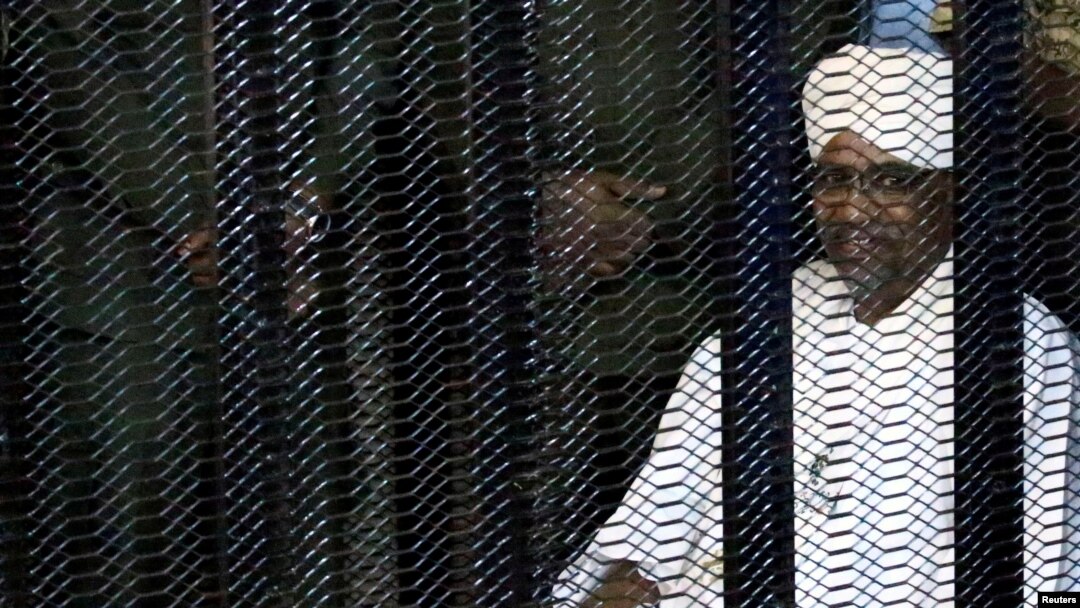 FILE - Sudan's former president Omar Hassan al-Bashir sits guarded inside a cage at the courthouse where he is facing corruption charges, in Khartoum, Aug. 19, 2019.