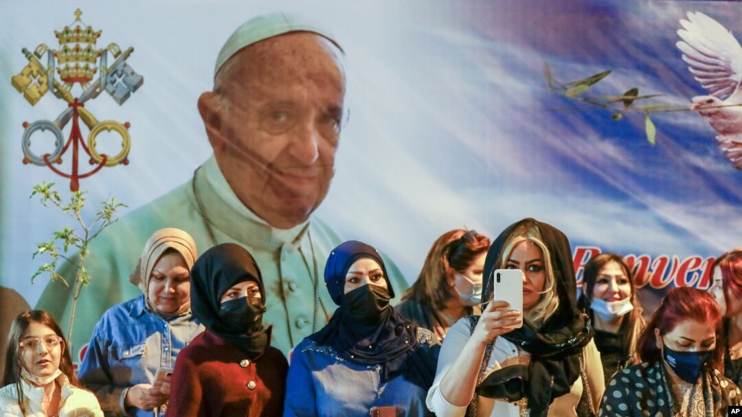Women wait outside the Chaldean Cathedral of Saint Joseph, in Baghdad, Iraq, March 6, 2021, where Pope Francis, shown on a giant poster in the backround, officiated mass. 