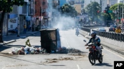 Un motociclista pasa al lado de un contenedor de basura quemado durante las protestas populares de este lunes contra el resultado oficial de la elección presidencial en Venezuela, en Caracas, la mañana del martes 30 de julio de 2024.