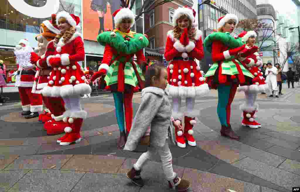 A girl walks in front of performers clad in Santa Claus outfits during a street event to promote Christmas season at a shopping district in Seoul, South Korea.