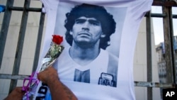 FILE - A man places a flower on a jersey with the face of the late football star Diego Maradona during a march to demand answers regarding his death in Buenos Aires, Argentina, March 10, 2021. 