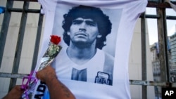 FILE - A man places a flower on a jersey with the face of the late football star Diego Maradona during a march to demand answers regarding his death, in Buenos Aires, Argentina, March 10, 2021. 