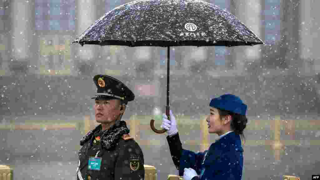 中国人大会议期间，人民大会堂外面，一位女服务员摆拍照片，在降雪时为武警打伞。（2018年3月17日）